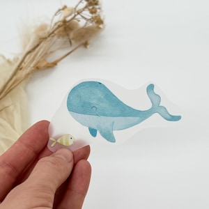Könnte beinhalten: Ein transparenter Aufkleber mit einem blauen Wal und einem gelben Fisch. Der Wal schwimmt nach rechts und der Fisch nach links. Der Aufkleber befindet sich auf einem weißen Hintergrund.