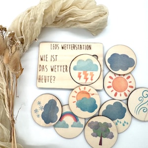 May include: Wooden weather station toy with the text "LEOS WETTERSTATION WIE IST DAS WETTER HEUTE?" includes a wooden board with a cutout for weather symbols. Round wooden tokens depict weather elements like sun, clouds, rain, and rainbow.