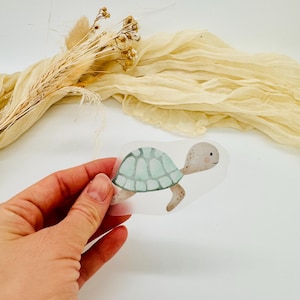 Könnte beinhalten: Ein Aufkleber im Aquarellstil einer Schildkröte mit hellblauem Panzer und beigem Körper. Die Schildkröte wird von einer Hand vor einem weißen Hintergrund mit beigem Stoff und Trockenblumen gehalten.