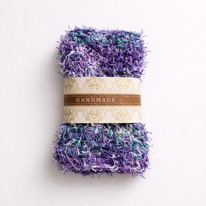 Puede incluir: Un estropajo rectangular hecho a mano en tonos morados, verde azulado y blanco. El estropajo está envuelto con una banda marrón con la palabra "HANDMADE" en letras doradas. La textura parece suave y en bucle.