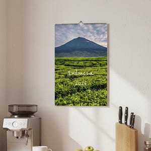 May include: A 2026 wall calendar depicting an Indonesian landscape. The image shows a green tea plantation with a mountain under a cloudy sky. The calendar is hanging on a white wall.