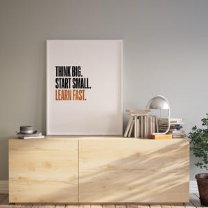 Puede incluir: Una impresión enmarcada en blanco con el texto "Think Big. Start Small. Learn Fast." en texto negro y naranja. La impresión está colgada en una pared gris claro sobre un armario de madera clara.
