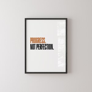 May include: A framed print with the words "PROGRESS. NOT PERFECTION." in a bold, modern font. The word "PROGRESS" is in a warm, burnt orange color, while "NOT PERFECTION" is in black. The frame is black, and the background is white.