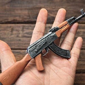 以下が含まれることがあります： 木製の銃床とハンドガード、黒いマガジンを備えた、AK-47ライフルのミニチュアレプリカ。 モデルは手で持たれており、そのサイズと詳細を示しています。 バレルとレシーバーは金属製です。