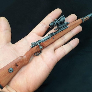 Op de afbeelding: Een miniatuur geweer met een vizier, met een houten kolf en metalen componenten. Het geweer is bruin en zilverkleurig, met een gedetailleerd ontwerp. De algehele esthetiek suggereert een verzamelobject of model.