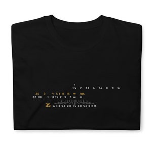 以下が含まれることがあります： カメラの絞り値のスケールが黄色で描かれた黒いTシャツ。絞り値と距離がフィートとメートルで表示されています。スケールは次のとおりです。1.4、2、2.8、4、5.6、8、11、16、2.5、3、4、5、6、8、15、∞、0.7、0.8、1、1.2、1.5、2、3、7、∞、35、16、11、8、5.6、2.8、1.4、2.8、5.6、8、11、16.