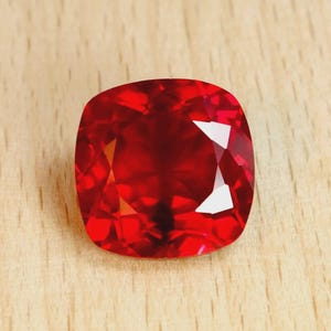 Peut inclure: Une pierre précieuse rubis taillée en coussin. La gemme est d'un rouge profond et vibrant, avec de multiples facettes réfléchissant la lumière. Le rubis est posé sur une surface en bois clair.