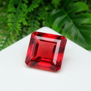 Puede incluir: Una gema roja, tallada en forma cuadrada, con una superficie facetada. La gema está colocada sobre una superficie blanca, con follaje verde en el fondo. El color de la gema es un rojo intenso y rico.