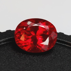 Peut inclure: Une pierre précieuse rouge vif, taillée en forme ovale et à facettes. La gemme présente de multiples surfaces réfléchissantes, créant un jeu de lumière brillant. La pierre est posée sur un fond noir, soulignant sa couleur vive et sa clarté. Idéale pour la joaillerie.