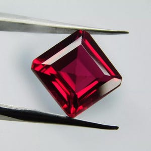 Op de afbeelding: Een dieprode vierkante edelsteen met een facetgeslepen vorm. De edelsteen wordt vastgehouden door een pincet.