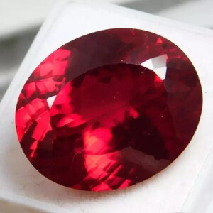 May include: A faceted, oval-shaped gemstone with a deep red hue. The gem is set against a white background, showcasing its brilliant cut and internal reflections. The polished surface of the stone catches the light, highlighting its rich color and clarity.