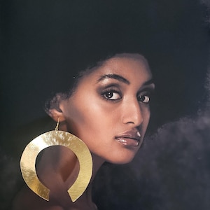 Peut inclure: Portrait d'une personne aux cheveux et à la peau foncés, portant de grandes boucles d'oreilles dorées. Les boucles d'oreilles ont un design unique, ouvert. Le fond est un dégradé sombre et flou.