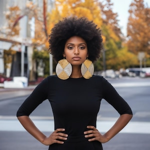 May include: A person wearing large, circular earrings with gold and silver geometric patterns. They are dressed in a black long-sleeved top with their hands on their hips. The background shows a blurred street scene with autumn foliage.