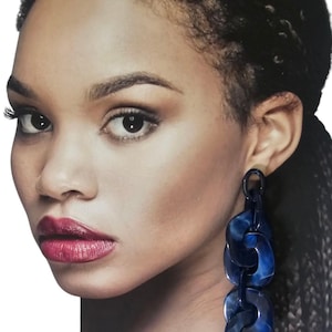 Puede incluir: Una mujer con cabello oscuro y aretes de cadena azul oscuro. Los aretes están hechos de un material veteado y cuelgan. La mujer tiene ojos oscuros y lápiz labial rojo.