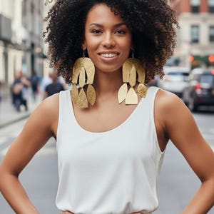 May include: A woman wearing large, gold-colored statement earrings. The earrings have a layered, geometric design. She is wearing a white tank top and standing on a city street with her hands on her hips.