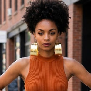 May include: A pair of large, curved, gold-toned metal hoop earrings with a hammered, textured finish. The earrings are wide and semi-circular in shape, featuring a polished, reflective surface that catches the light, worn as statement jewelry.