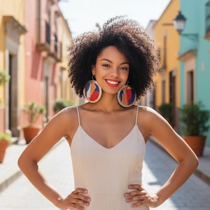 May include: A woman wearing large, round earrings with colorful abstract designs. She is smiling and wearing a cream-colored dress with spaghetti straps. The background features a colorful street scene with buildings in shades of yellow, pink, and blue.