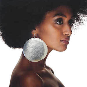 Puede incluir: Una mujer de piel oscura y cabello negro voluminoso lleva pendientes grandes, plateados y circulares. Los pendientes tienen una superficie texturizada y cuelgan de sencillos ganchos plateados. La imagen es un retrato con fondo blanco.