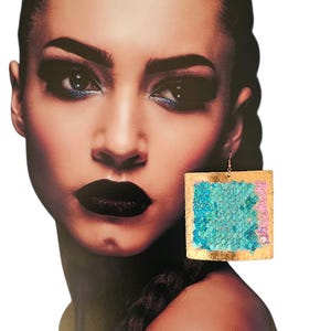 May include: Square sequin earrings with a gold border. The sequins are in shades of blue and pink. The earrings are hanging from a gold hook. The background is a portrait of a woman.