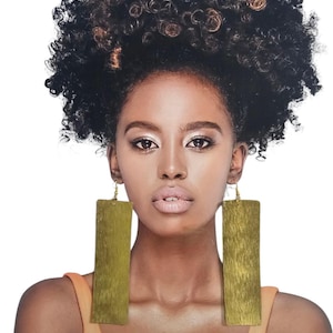Può includere: Una persona con capelli ricci scuri indossa grandi orecchini rettangolari dorati. Gli orecchini sono strutturati e pendono da piccoli ganci dorati. Indossa una canotta arancione chiaro.
