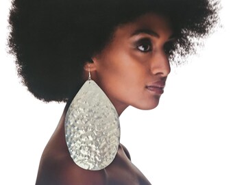 May include: Silver-colored, hammered metal teardrop earrings. The earrings are large and dangle from a silver hook. The earrings have a textured surface and are worn by a person with dark skin and black hair.