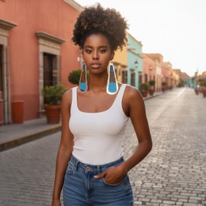 May include: A woman wearing large, geometric earrings with blue and brown accents. She is wearing a white tank top and blue jeans. The earrings are the focal point, set against a colorful street backdrop.