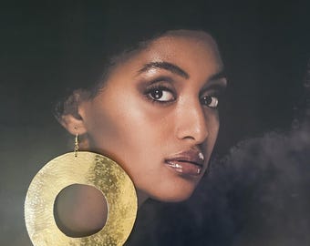 Pode incluir: Uma mulher de pele escura com grandes brincos de argola dourados. Os brincos são circulares com uma textura martelada. A mulher tem cabelo escuro e está olhando para a câmera. O fundo é escuro.