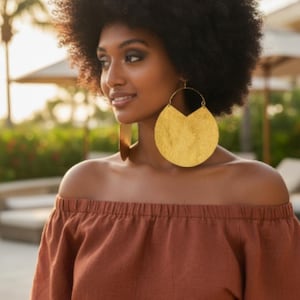 May include: A woman wearing large, gold-colored, circular earrings. She is also wearing an off-the-shoulder, rust-colored top. Her hair is styled in a large afro. The background is blurred, with greenery and sunlight.