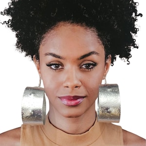 May include: A woman with dark curly hair and silver hoop earrings. The earrings are large, cylindrical, and have a textured surface. She is wearing a tan turtleneck top. The background is white.