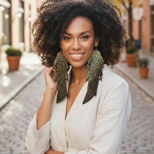 Puede incluir: Una mujer con pendientes grandes con estampado dorado y negro. Los pendientes tienen un diseño en capas, similar a plumas. Lleva una blazer color crema y sonríe. El fondo es una calle adoquinada.