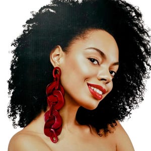 May include: A woman with dark curly hair and red lipstick smiles, wearing large, dangling red chain link earrings. The earrings are made of a translucent material, and the links are connected in a vertical line.