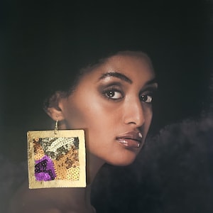 May include: A woman wearing a large, square gold earring. The earring is decorated with sequins in shades of purple, gold, and white. The woman has dark skin and is looking at the camera.