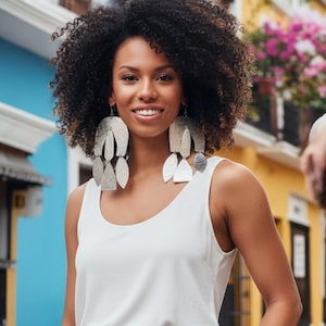 May include: A woman wearing large, silver-colored leaf-shaped earrings. The earrings are layered and dangle. She is wearing a white tank top. The background features colorful buildings.