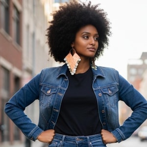 May include: A woman wearing a denim jacket, black t-shirt, and jeans. She is wearing large, peach-colored earrings with white accents. The background is a blurred city street.