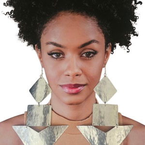 May include: A woman wearing large, silver-toned geometric earrings. The earrings feature a diamond shape at the top, a square in the middle, and a triangle at the bottom. The earrings have a hammered texture and hang from a silver chain.