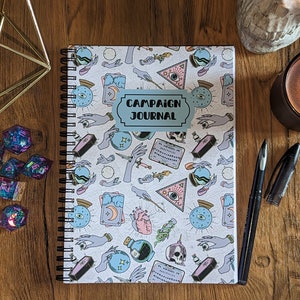 May include: A white spiral bound journal with a colourful pattern of hands, eyes, skulls, and other mystical symbols. The cover has a blue sticker with the text "Campaign Journal".