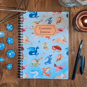 May include: A blue notebook with a spiral binding and a dragon pattern. The cover says "Campaign Journal". There are blue dice and a pen on a wooden surface.