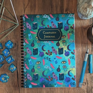 May include: A blue and green spiral bound journal with a black title that reads "Campaign Journal". The cover features a repeating pattern of various illustrations including eyes, hands, feathers, potions, and books. The journal is on a wooden table with dice and a pen.