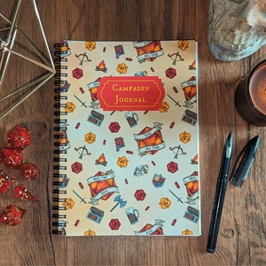 May include: A spiral bound notebook with a white cover and a colorful pattern of dice, swords, and other fantasy-themed illustrations. The cover has the text "Campaign Journal" in red and white.