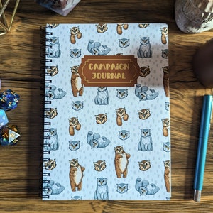 May include: A blue spiral bound notebook with a white cover featuring a repeating pattern of brown owls. The cover has the text "Campaign Journal" in gold lettering.
