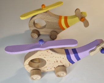 May include: Two wooden toy helicopters, one painted yellow and orange, the other painted purple and blue. Both have spinning propellers and wheels.