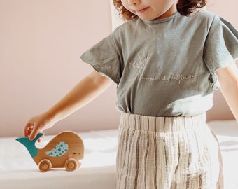 May include: A young child in a light blue t-shirt with the text "l'amour et des fleurs" embroidered on it, and white and brown striped pants, plays with a wooden toy whale with blue and white polka dots.