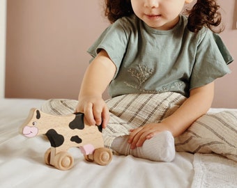 May include: A young child plays with a wooden toy cow with black and white spots. The cow has pink details and wheels. The child is wearing a light blue shirt with an embroidered design and striped pants.