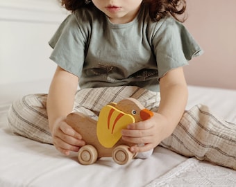 May include: A young child plays with a wooden toy animal. The toy is brown with yellow and orange accents and has wheels. The child is sitting on a white bed with a white and gray blanket.