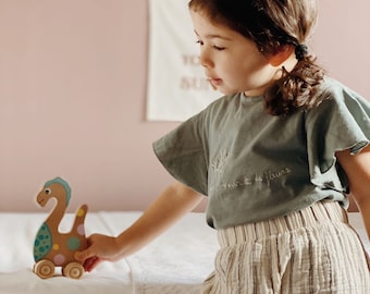 May include: A young child wearing a light blue shirt with the text "un jour de fleurs" embroidered on it and striped pants plays with a wooden toy dinosaur with colorful polka dots. The toy has wheels and is painted in shades of brown, blue, yellow, and green.