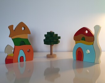 May include: Three wooden toy houses, one shaped like a snail, one shaped like a mushroom, and one shaped like a house with a green roof. A small green tree is in the center.