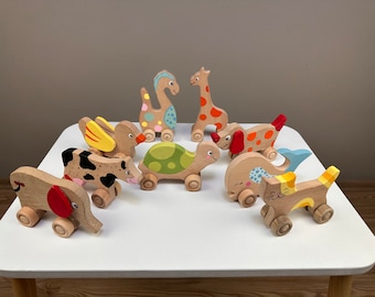 May include: A set of nine wooden pull toys featuring various animals, including a cow, a turtle, a giraffe, a cat, a duck, a whale, an elephant, and a dinosaur. The toys are painted in bright colors and have wheels on the bottom.
