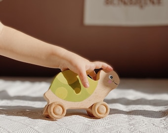May include: A wooden toy turtle with green and yellow painted shell and wheels. The turtle is on a white blanket.