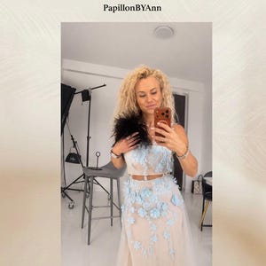 Może przedstawiać: Dwuczęściowy zestaw składający się z bezrękawnika typu crop top oraz spódnicy do ziemi w jasnobeżowym odcieniu. Ubiór ozdobiony jest jasnoniebieskimi aplikacjami kwiatowymi oraz czarnym wykończeniem z piór przy dekolcie. Widoczny jest napis PapillonBYAnn.