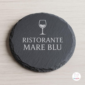 Peut inclure: Un dessous de verre rond en ardoise gris foncé. Il présente le texte "RISTORANTE MARE BLU" et un dessin de verre à vin blanc. Le dessous de verre a un bord naturel et irrégulier, idéal pour les boissons.